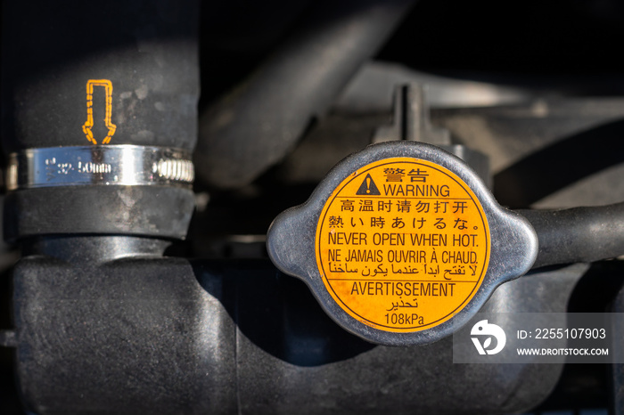 Orange radiator cap with warning labels in different languages: never open when hot, and blurred car radiator hose with yellow dotted arrow under the open hood of a car. Closeup view