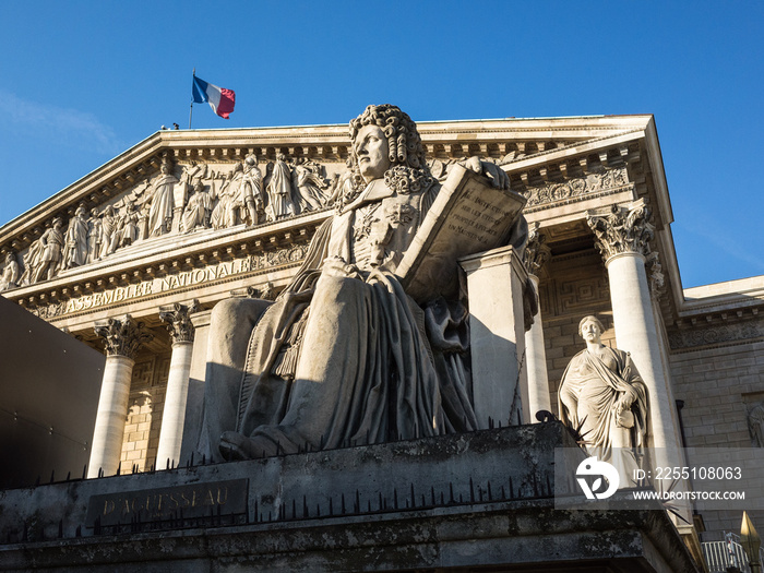 Facade with colonnade of the Palais Bourbon in Paris, seat of the french National Assembly, with the statue of Francois d’Aguesseau and the french flag flying on the roof against blue sky.