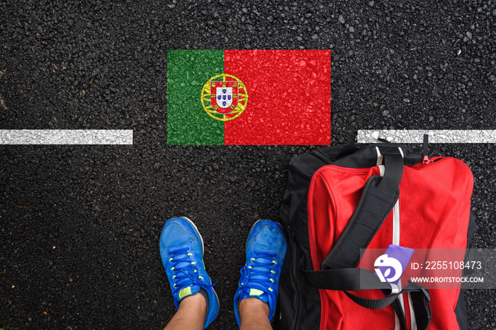 a man with a shoes and travel bag is standing on asphalt next to flag of Portugal and border