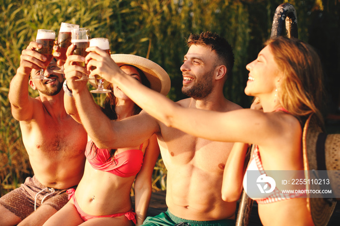 Group of young people toasts with glasses of beer by the river during the summer sunny day