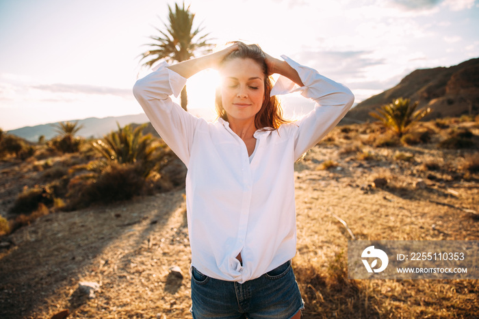 Portrait of an attractive young woman posing smiling in a wonderful desert landscape with a palm tree and mountains in the background.  Advertising photography. Fashion and lifestyle concept