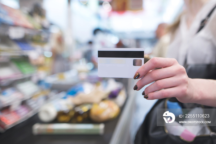 Hand with a white credit card on the background of a supermarket cash desk. Credit card in a woman’s hand when paying in a supermarket.