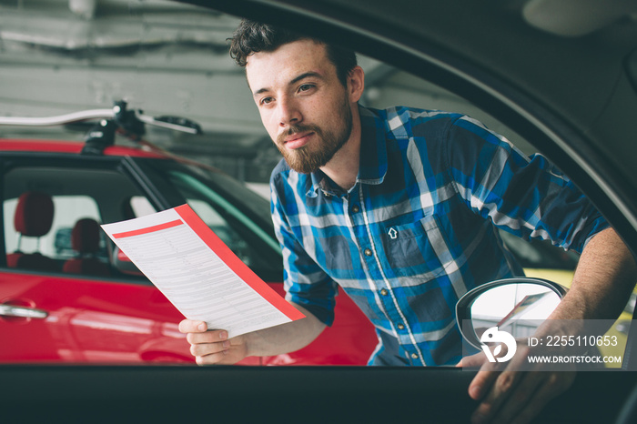 Perfect lines. The young dark-haired bearded man examining car at the dealership and making his choice. Horizontal portrait of a young guy at the car. He is thinking if he should buy it