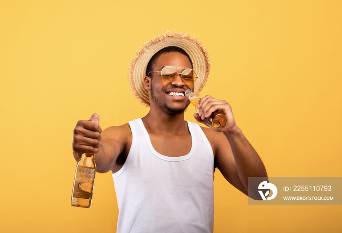 Positive black guy in summer outfit holding two bottles of beer, drinking alcoholic beverage on yellow studio background