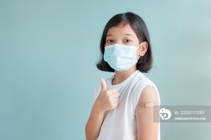 Asian little girl wearing face mask vaccinated Gesturing Thumbs Up  Showing arm bandage to protect COVID-19 spread on blue background