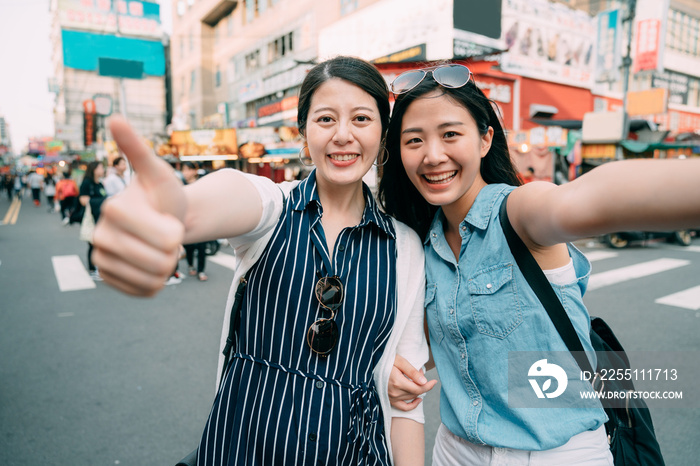 two happy asian tourist girls are smiling at the camera with thumb up gesture while taking a selfie on the street of a sightseeing shopping area in summer.