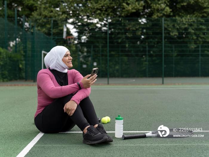 UK,Sutton,Woman in headscarf sitting on tennis court,holding smart phone