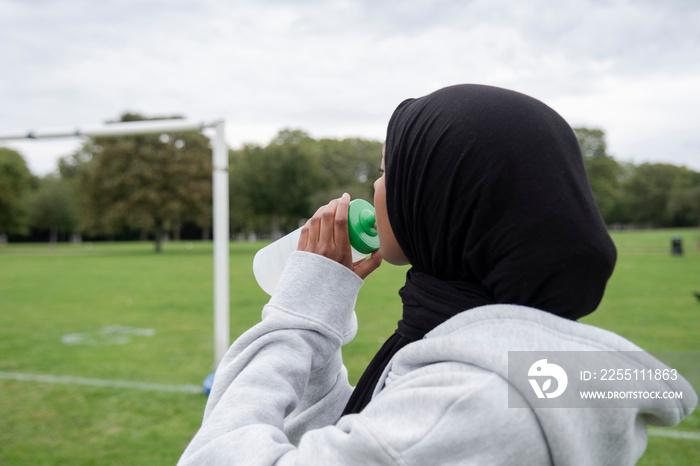 Woman in hijab drinking water from bottle in soccer field