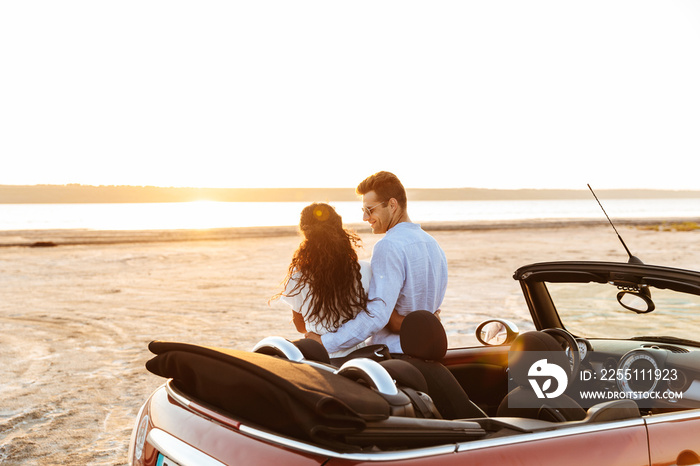 Photo from back of attractive multiethnic couple hugging together while standing by car on beach