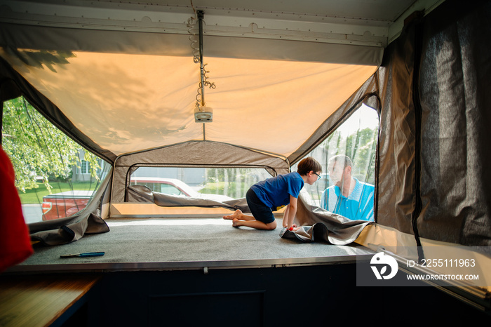 A boy inside of a pop-up camper peeks out of one of the windows at his dad