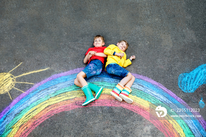 two school kids boys having fun with with rainbow picture drawing with colorful chalks on asphalt. Siblings, twins and best friends in rubber boots painting on ground playing together.