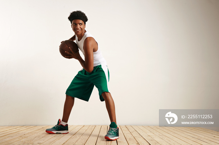 Full length horizontal shot of a young attractive African American with afro and headband playing happily with a vintage leather basketball against white wall and light wooden floor.