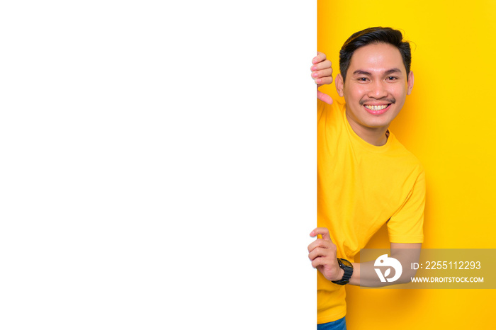 Cheerful young Asian man in casual t-shirt peeping out the side of white advertisement board isolated on yellow background. Promotion billboard concept