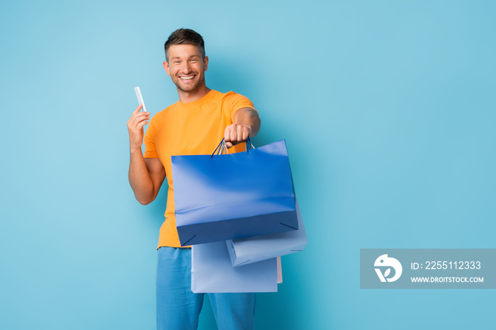 happy man holding shopping bags and smartphone on blue