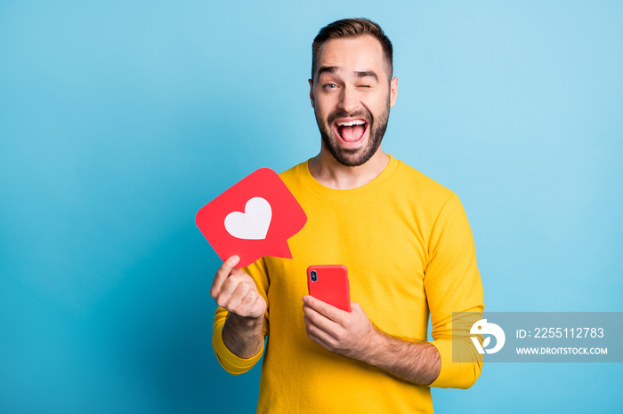 Photo of young happy excited smiling cheerful man wink eye hold phone and heart icon isolated on blue color background