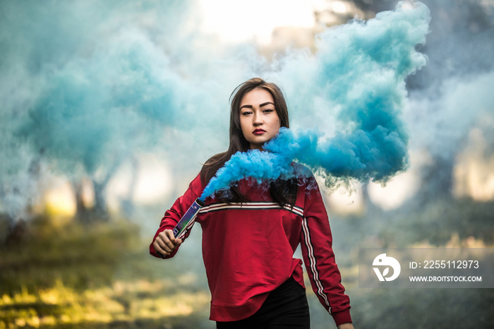 Young asian woman holding blue colorful smoke bomb on the outdoor park. Blue smoke spreading