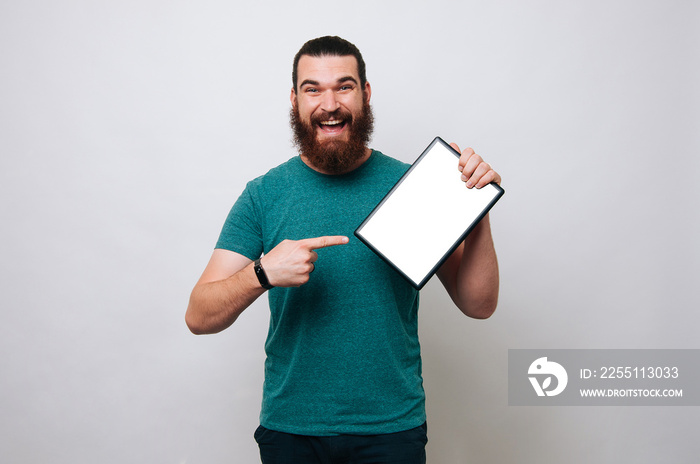 Cheerful young bearded man pointing on white screen tablet over grey background