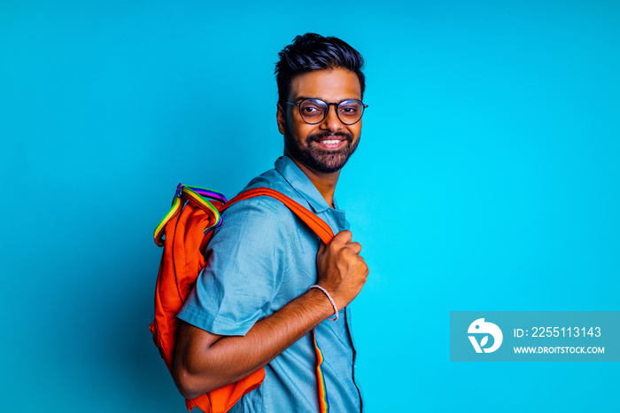 handsome young bearbed indian man with eye glasses in blue cotton t-shirt with orange rainbow backpack in studio background