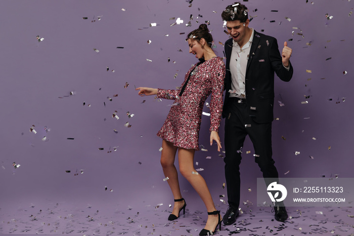 Young brunette in mini glitter dress and shoes smiling and dancing with attractive man wearing black suit and white shirt. Pair posing with confetti against violet background