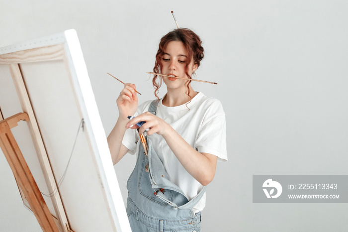 Young woman artist working in the workshop, squeezing paints on her brush. Work with oil paints, brushes and easel. Creative career