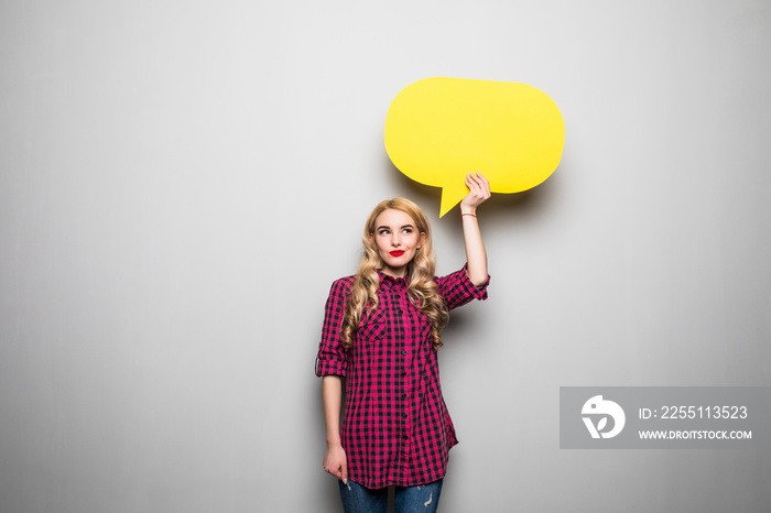 Beautiful blond young woman holding yellow blank speech bubble over grey background