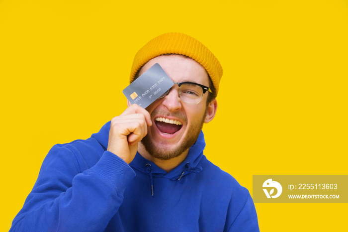 Happy young caucasian guy with credit, debit card in hand smiling cover his eye ready to spend money. Young man in blue hoodie with mockup bank card isolated on yellow background. Shopping concept