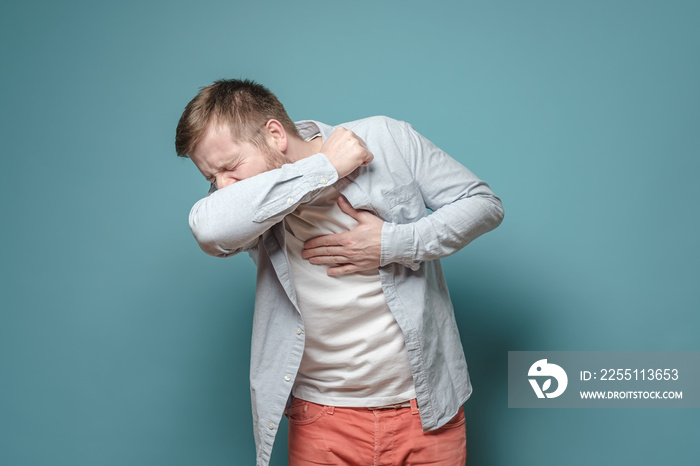 Young European man coughs at elbow and holds hand on chest, experiencing pain. Concept of the spread of the virus.