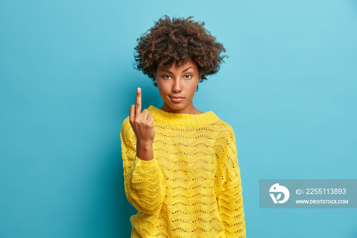 Serious young pretty Afro American woman shows middle finger bad sign asks not to bother her wears yellow sweater isolated on blue background. Body language concept. Provocation and rude attitude