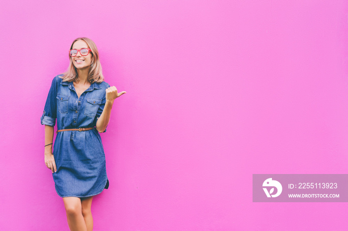 Copy space for advertisement. Colorful portrait of happy smiling young woman pointing away against pink wall.