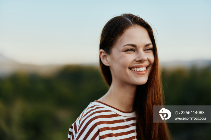 Close-up portrait of a young woman with a beautiful smile with teeth in a striped t-shirt against the background of trees