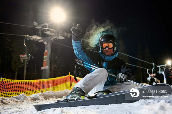 Male skier in winter ski jacket and helmet holding ski poles and showing rock and roll gesture. Young man in ski goggles sitting on snow-covered slope. Concept of night skiing at ski resort.