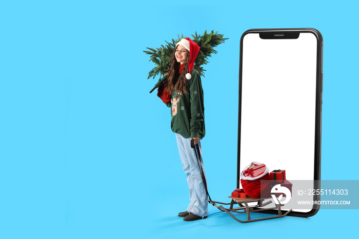 Beautiful girl in Santa hat with Christmas tree, sledge, presents and big smartphone on light blue background