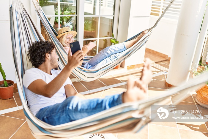 Young couple smiling happy relaxed using smartphone and reading book. Lying on hammock at terrace.