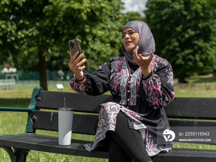 UK,Sutton,Woman in headscarf sitting on bench in park,talking on smart phone