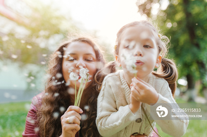 Young pretty women with small girl blowing dandelions in the park. Spend happy time together outside.