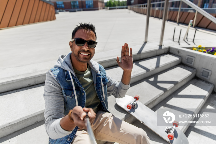 leisure, people and lifestyle concept - smiling indian man in sunglasses waving hand and taking picture by selfie stick on roof top