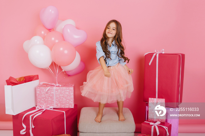 Charming excited little girl in tulle skirt having fun, dancing on chair suround birthday presents, balloons isolated on pink background. Expressing true positive emotions, happiness of childhood