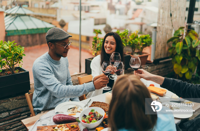 Long awaited meeting of best friends on summer rooftop with cozy garden, Multicultural group of people making cheers with red wine glasses laughing and joking while celebrating meeting reunion outside