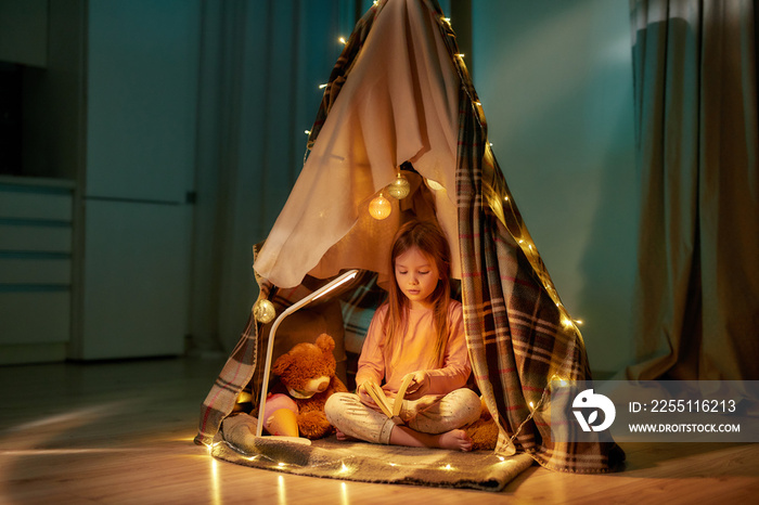 A cute girl wearing pyjamas sitting on a floor cross-legged barefoot in a self-made hut made of a plaid holding and scrolling a book with fancy garlands all around the hut