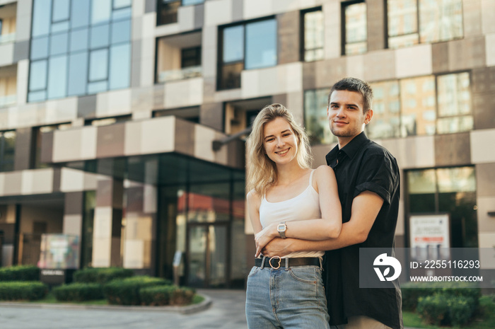 Street photo beautiful stylish young couple hugging against the backdrop of a modern building, looking in camera and smiling. Happy young family stands on the background of a modern apartment building