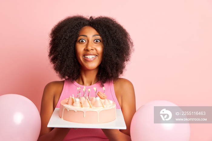 birthday woman holds pink cake with candles and staring with surprise and happy smile, isolated next to pink wall