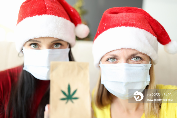 Two women in santa claus hats and protective masks on faces holding bag of marijuana. Legal sale and purchase of cannabis concept