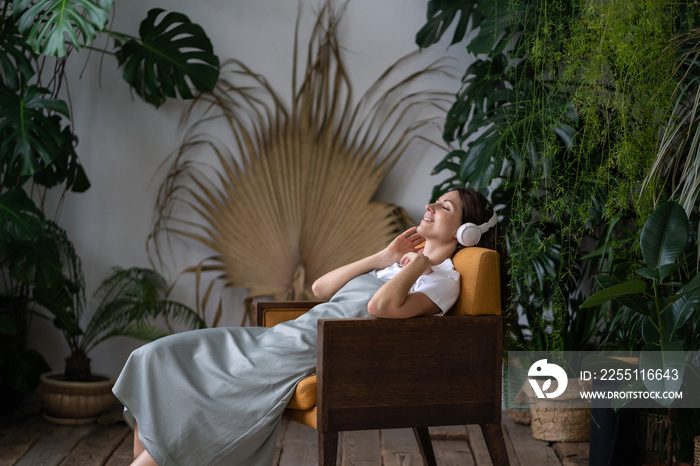 Relaxed girl lies surrounded by exotic green plants in the city botanical garden in wireless headphones. Smiling woman resting after working day listening to music, interesting podcasts in earphones.