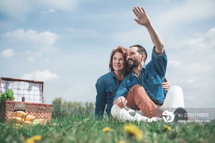 Hello. Cheerful mature man is waving hand to someone while sitting on blanket with his wife. Woman is embracing man and smiling. They are having picnic on the meadow