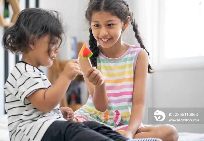 Young girl and little boy have fun eating sweet candy. Happy children holding sugar taffy enjoy delicious snacks. Cheerful sister playing with brother kid toffee in hand. Childhood enjoyment lifestyle