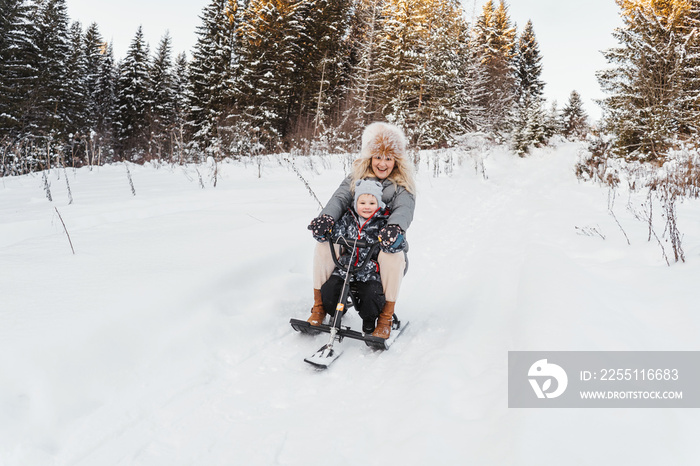 In winter, happy mom and son ride a snow scooter from the mountain in white snow. Sunny forest on background.