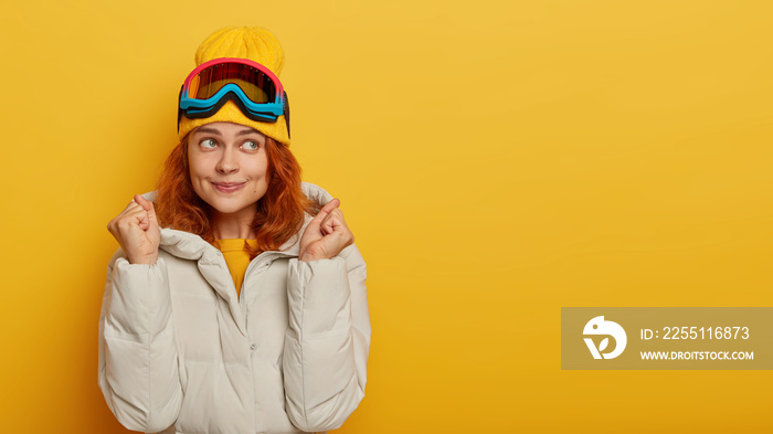 Thoughtful female tourist recreats during winter time, raises fists, anticipates for miracle, wears ski goggles, winter warm coat, going to try snowboarding first time, isolated over yellow background
