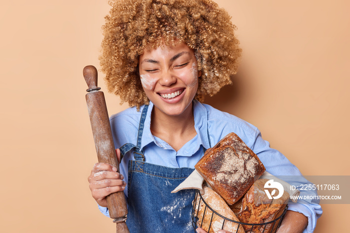 Horizontal shot of positive female baker holds rolling pin and baset full of freshly baked bread works in bakery shop wears blue shirt and denim sarafan enjoys eating organic products poses indoor