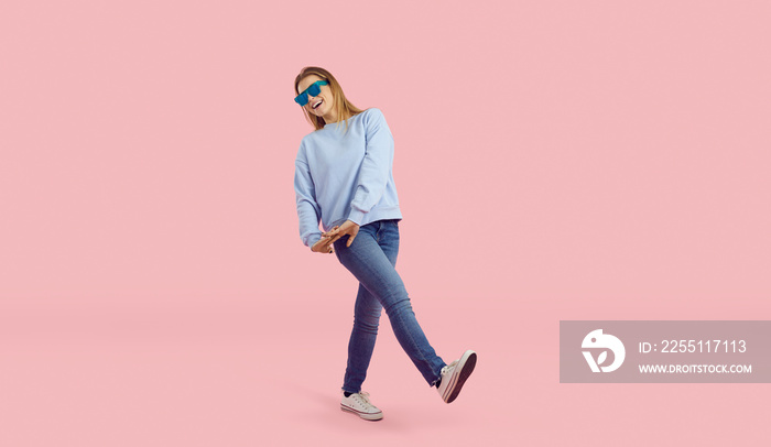 Happy funny young woman walking against a pastel pink colour studio background. Full length studio shot of a cheerful smiling young girl wearing trendy blue sweatshirt, jeans and crazy sunglasses