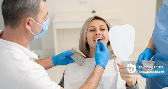 Dentist in blue medical gloves holds a sample of tooth enamel near a female patient with white teeth to pick up the right shade, teeth whitening procedure,